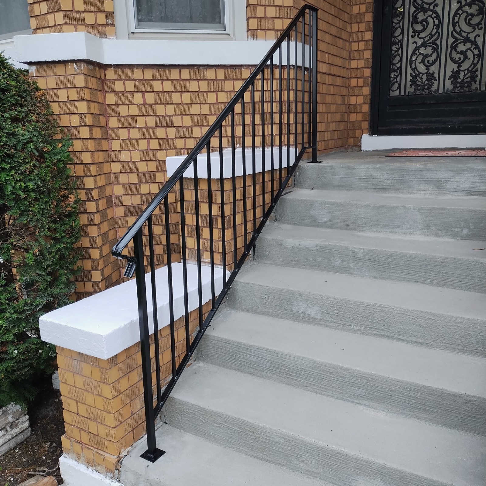 Simple black handrail installed along concrete front stairs at a brick home.